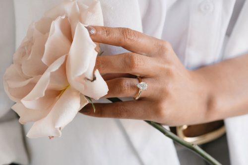 Iris engagement ring in yellow gold, worn on hand holding a pale pink rose, featuring a pear-cut diamond with petal-style prongs.