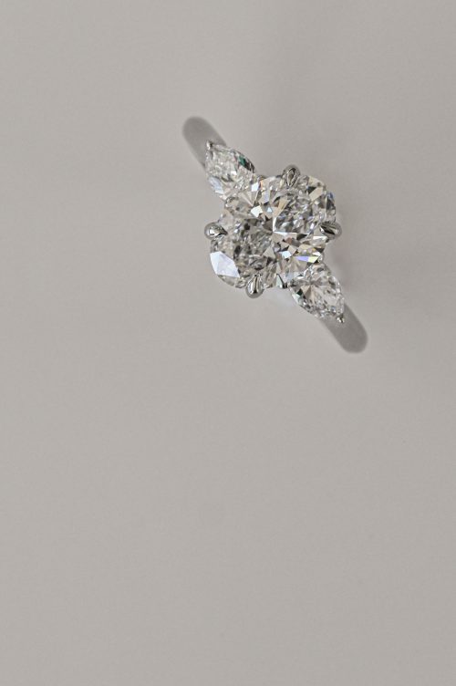 Rae three-stone ring in 19k white gold with yellow gold lock detail, featuring an oval center diamond with pear-shaped side stones.