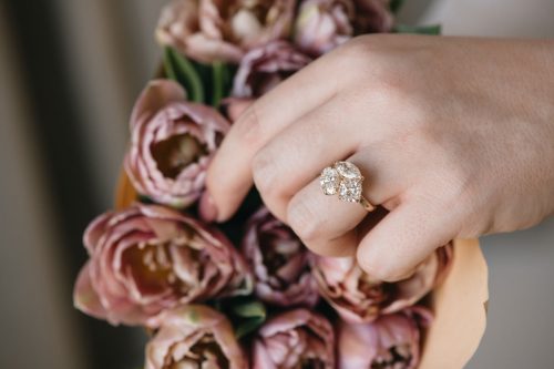 Three-stone diamond cluster ring in yellow gold, worn on hand holding soft pink tulips.