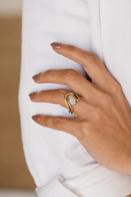 Mira engagement ring in 14k yellow gold, worn with the coordinating Mira wedding band, both featuring fluid, sculptural curves that interlock around the oval diamond in a seamless, modern embrace.