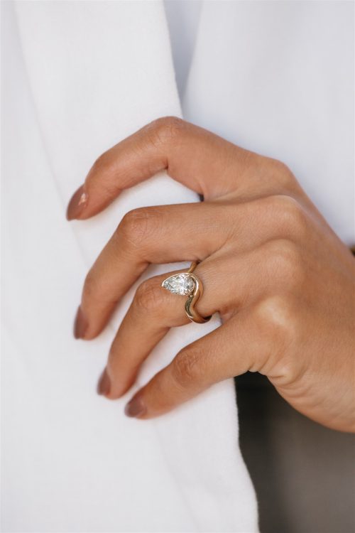 A close-up of a hand adorned with the Amara ring, showcasing a pear-shaped diamond embraced by a modern wave-shaped yellow gold band, reflecting a minimalist yet sculptural elegance against a soft white fabric backdrop.