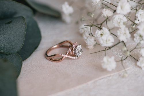 The Angelica ring rests delicately between baby's breath and eucalyptus, featuring a brilliant round diamond embraced by a split shank of rose gold—adorned with shimmering pink pavé diamonds that gracefully twist around the center stone.