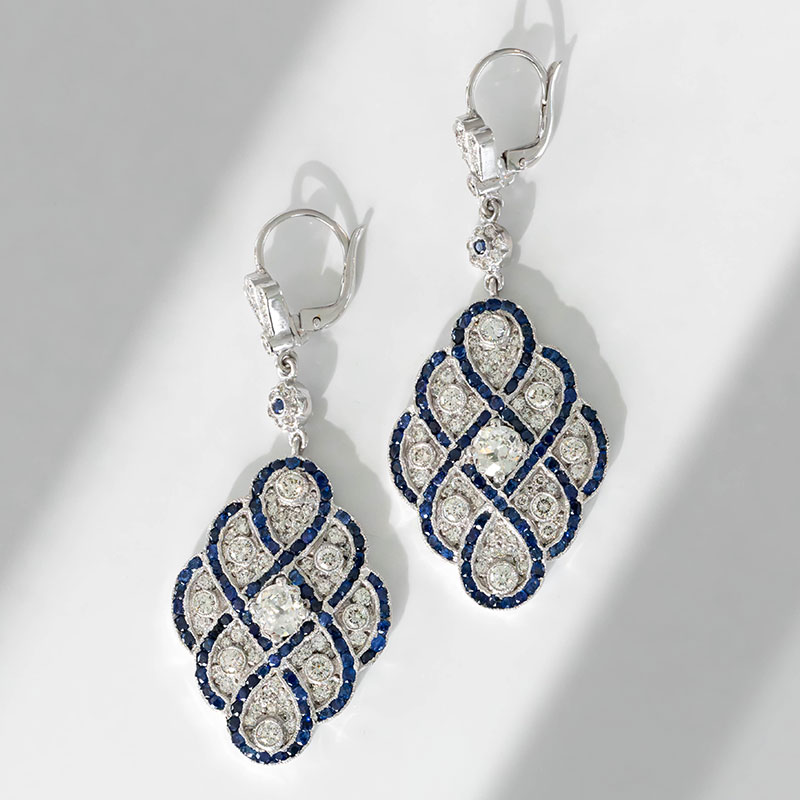 Art Deco-style drop earrings in white gold, featuring diamond clusters framed by blue sapphire borders in an intricate open lattice pattern.