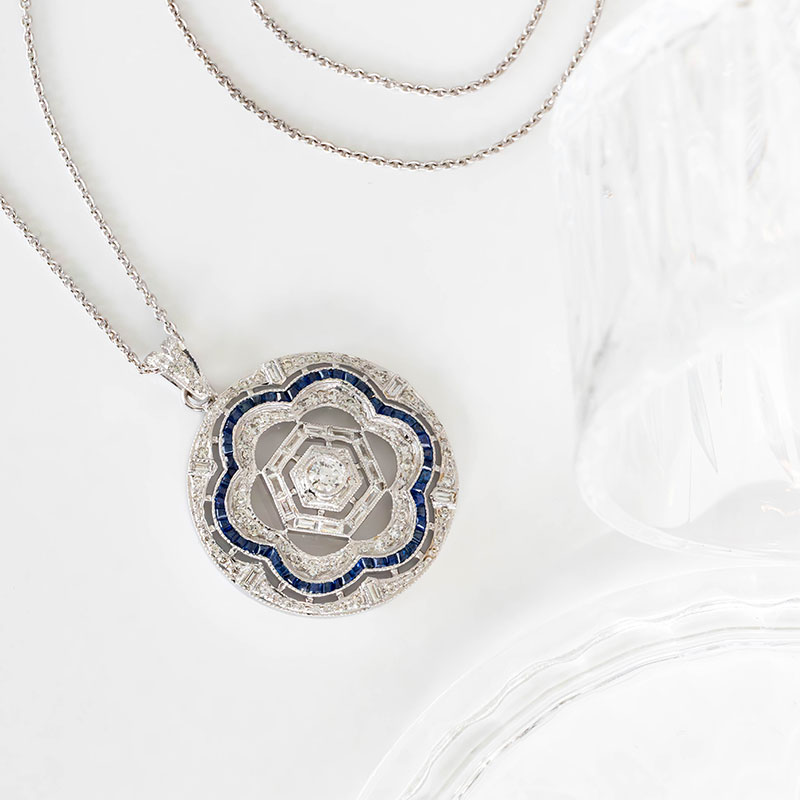 Round Art Deco pendant in white gold, featuring a geometric floral motif set with diamonds and framed by a blue sapphire border.