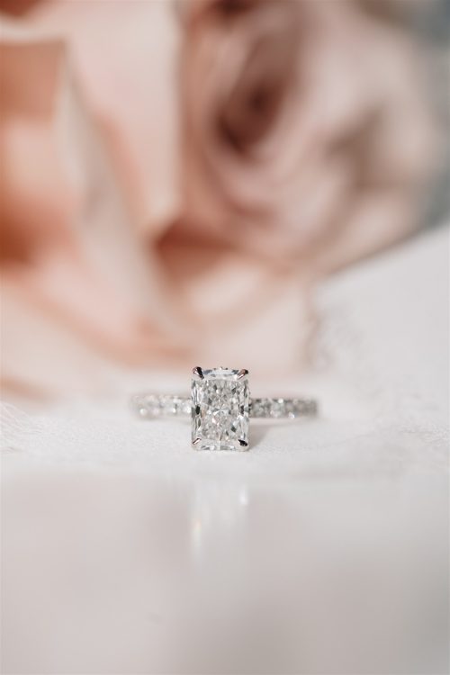 Front-facing view of the Cecelia engagement ring, showcasing a radiant-cut diamond set in a platinum band with delicate pavé detailing, softly styled against blush-toned florals.