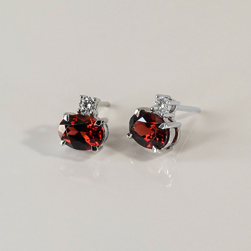 Oval-cut garnet stud earrings in white gold, each accented with a small round diamond above the main stone.