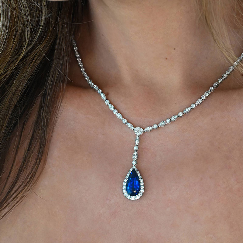 White gold necklace with a pear-shaped blue sapphire pendant surrounded by a diamond halo and accented by a diamond station chain.