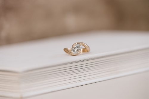 A close-up of the Selene ring displayed on a white book, featuring an oval-cut diamond gracefully cradled by a sculptural bypass-style yellow gold band.