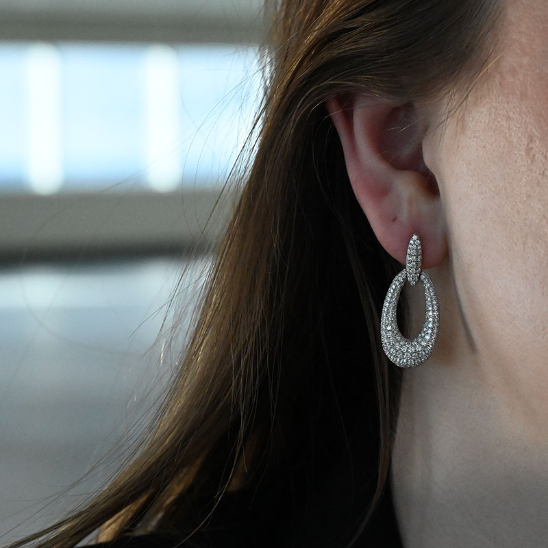 Close-up of a woman's ear wearing pavé diamond oval drop earrings with a small loop connecting to a larger open oval.